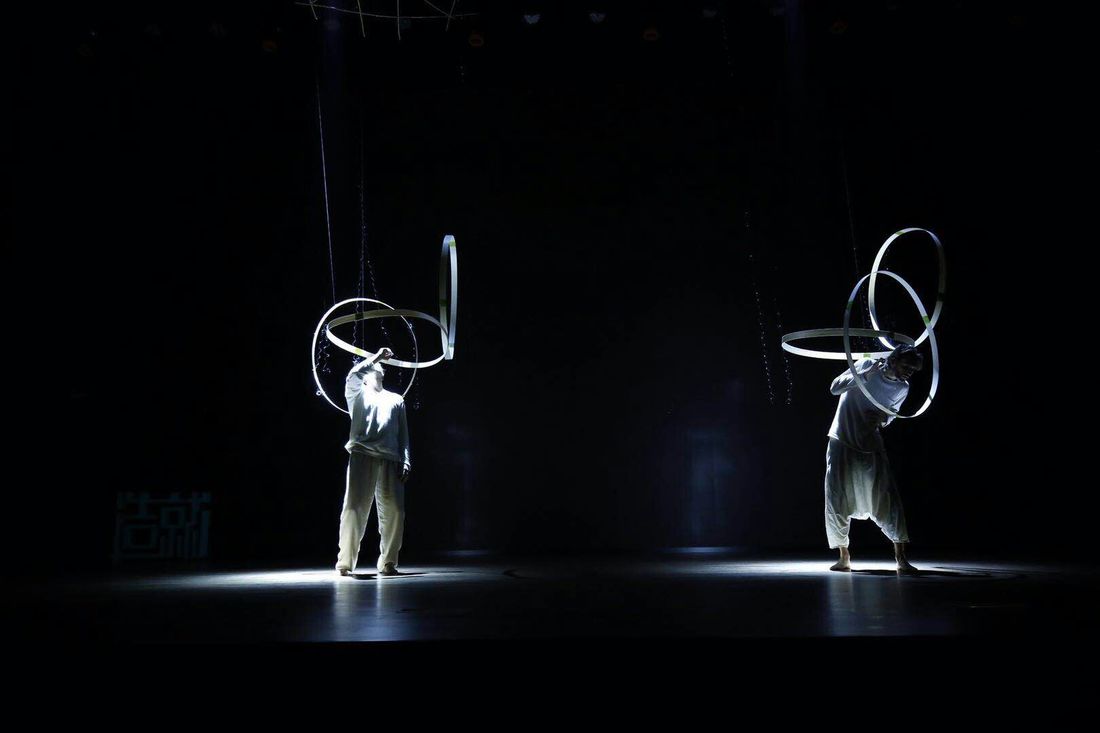Two dancers in white stand apart on a dark stage, each looking up into a cluster of suspended white rings.