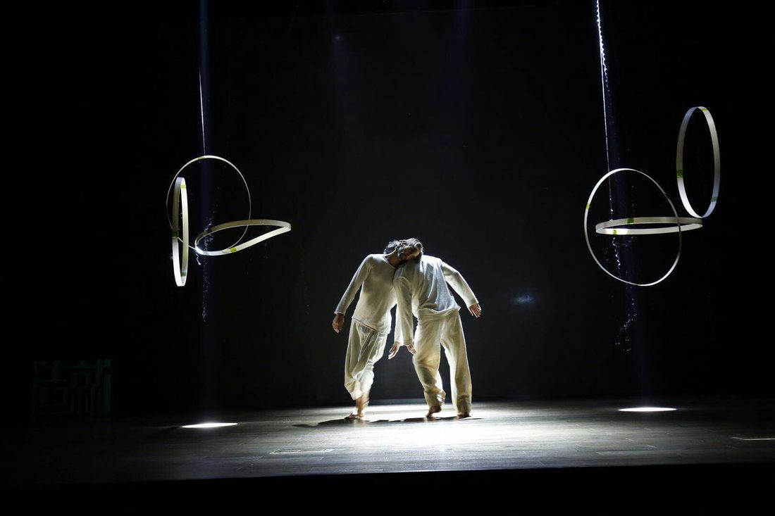 Two dancers in white share weight through their heads on a dark stage, suspended white rings hanging on either side.