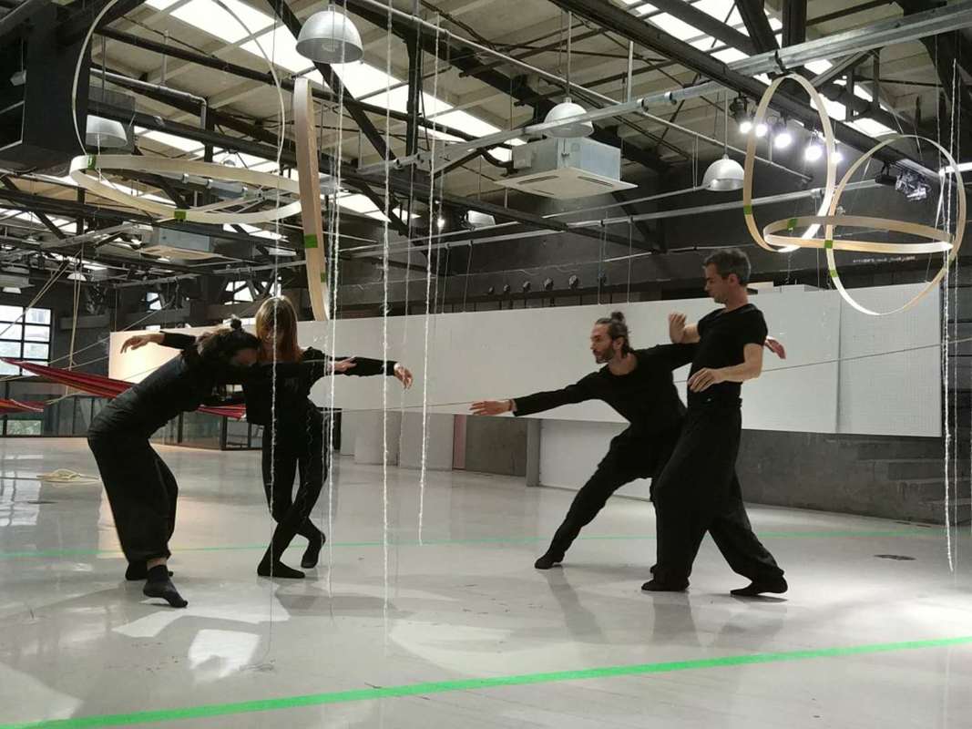 Four dancers in black rehearse among hanging ring sculptures in a bright industrial hall.