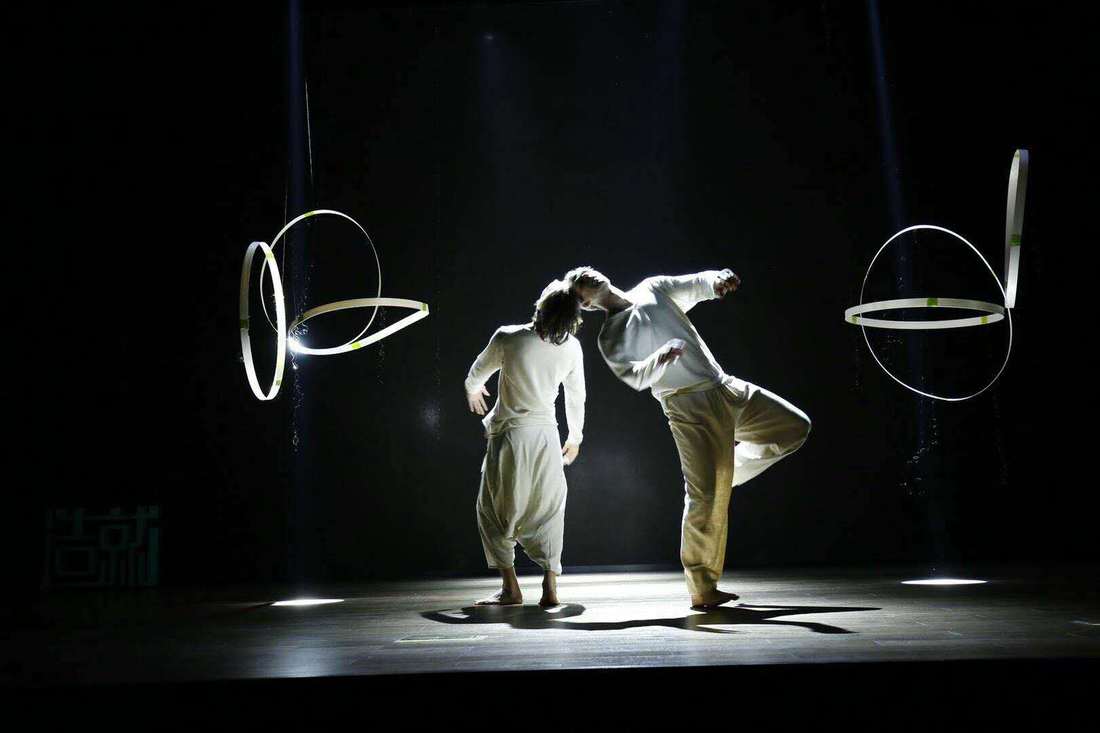 Two dancers in white lean into each other on a dark stage, suspended white rings on either side.