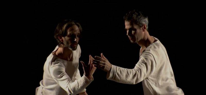 Two dancers reach toward each other, hands almost touching, against a black background.