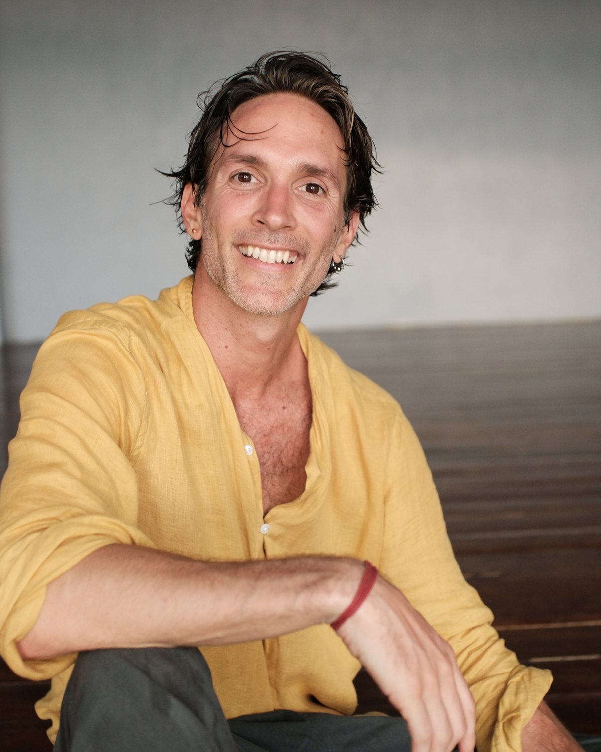 Eric Alessi sitting on a wooden studio floor in a yellow linen shirt, smiling.