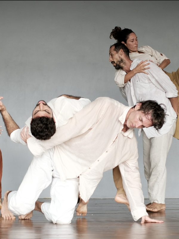 One dancer folded over the back of another in a studio with tall windows.