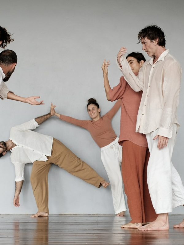 Five dancers in white and earth tones mid-lift against a grey wall.